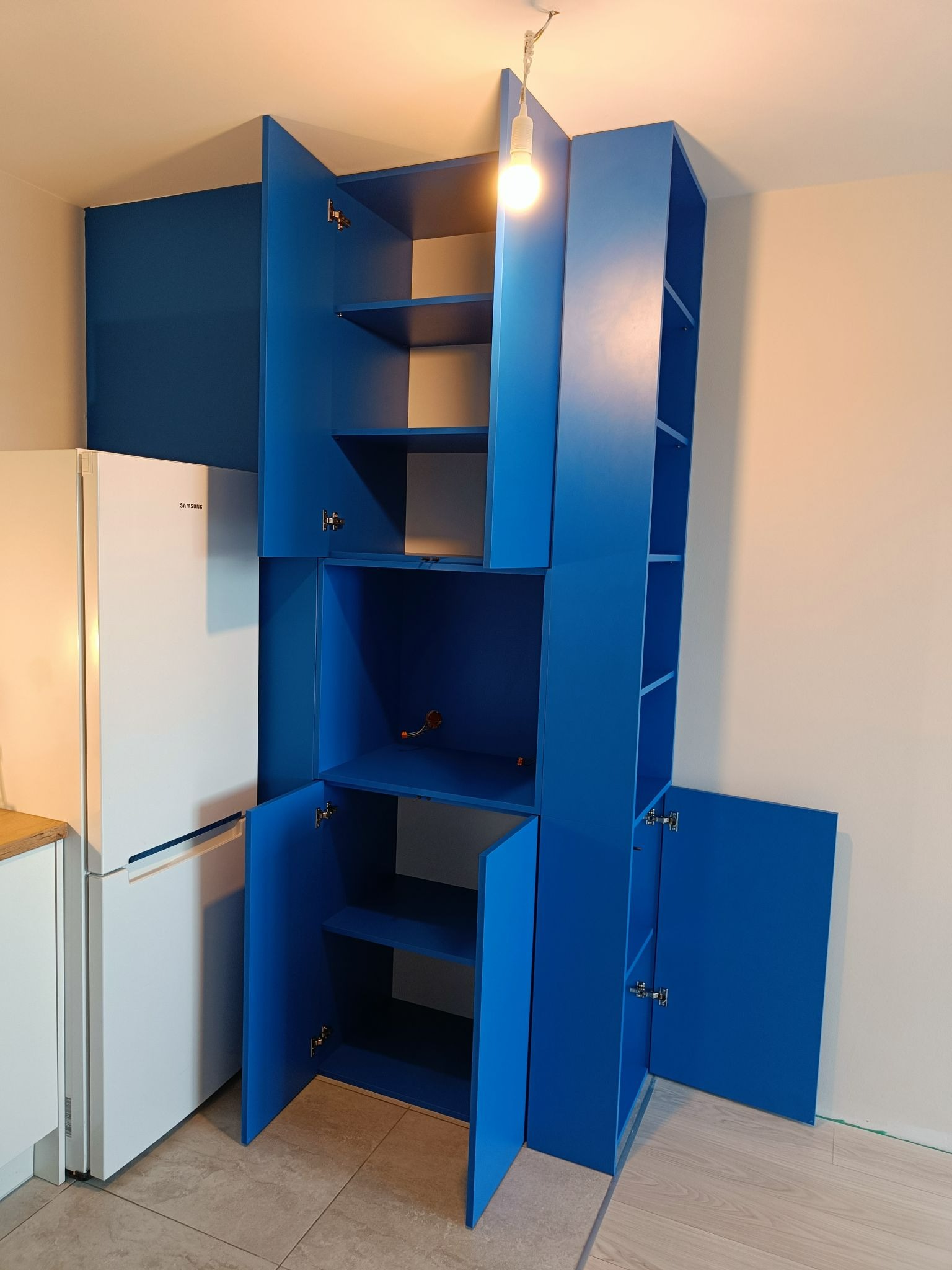 Wysoka, niebieska zabudowa meblowa z otwartymi szafkami w kuchni, obok biała lodówka Samsung, widoczna żarówka na suficie.