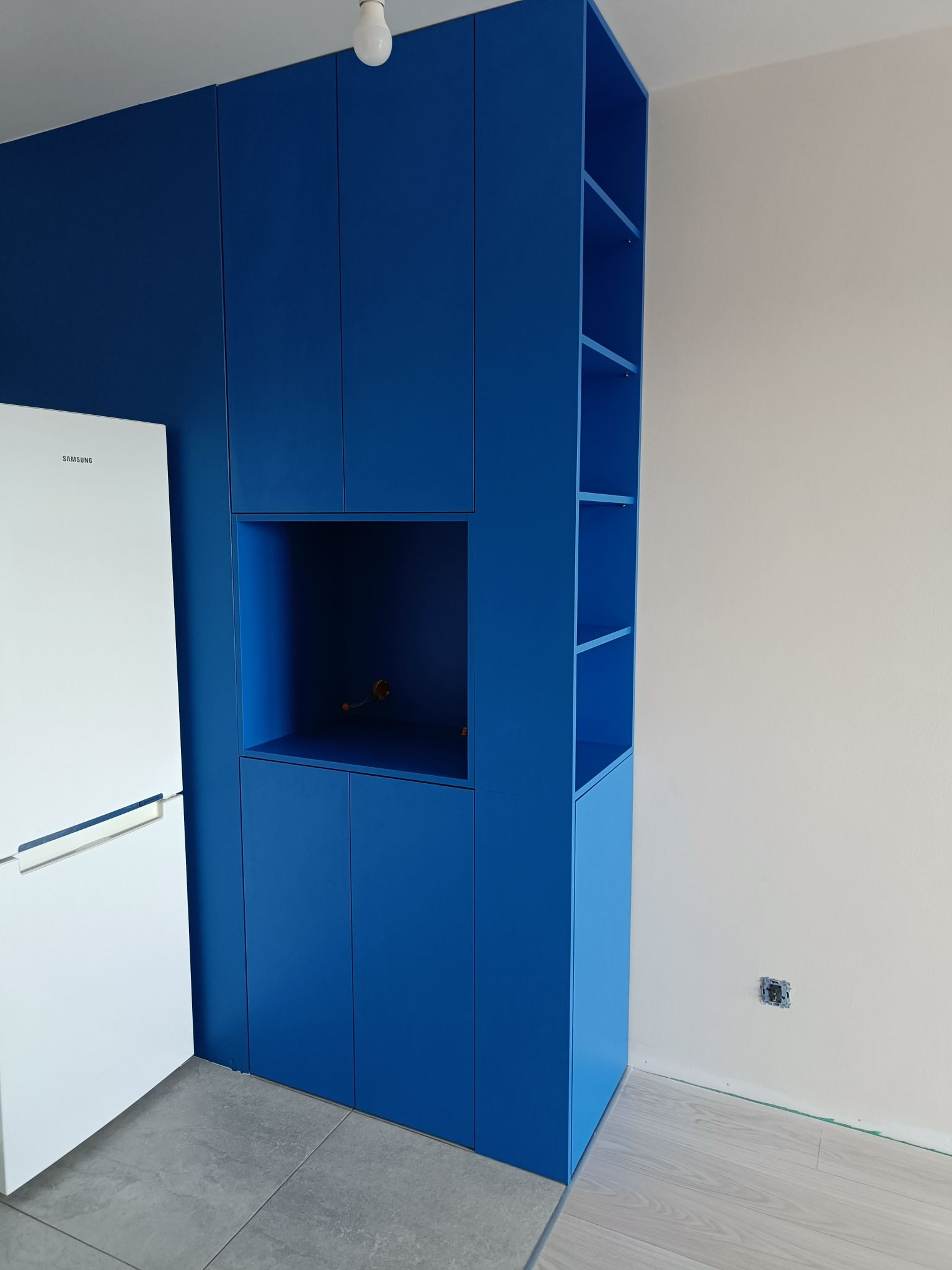 Niebieska zabudowa meblowa w kuchni z otwartymi półkami i miejscem na lodówkę, widoczne gniazdko elektryczne w ścianie.