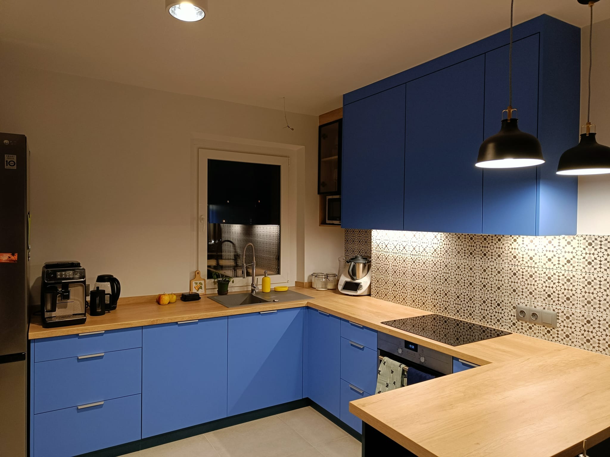 Niebieska zabudowa kuchenna z drewnianym blatem, widoczne sprzęty AGD, mozaika na ścianie, oświetlenie punktowe i wiszące lampy.