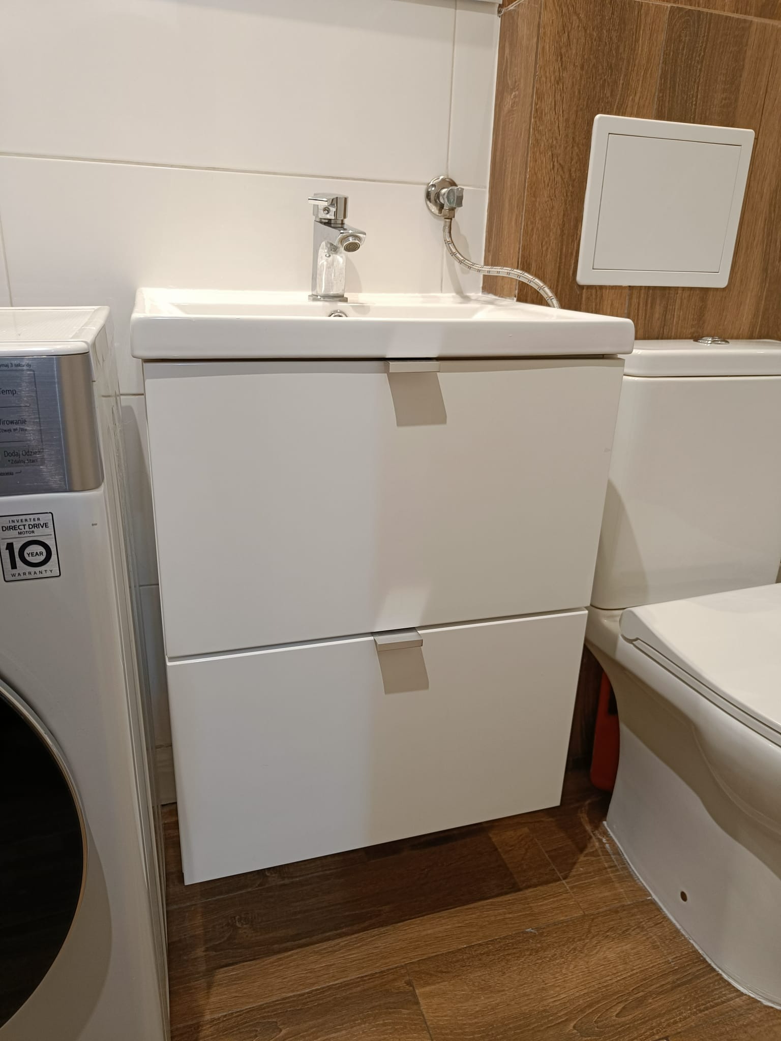 Biała umywalka z szafką podwieszaną w łazience, obok pralka i toaleta, widoczne drewnopodobne panele na ścianie.
