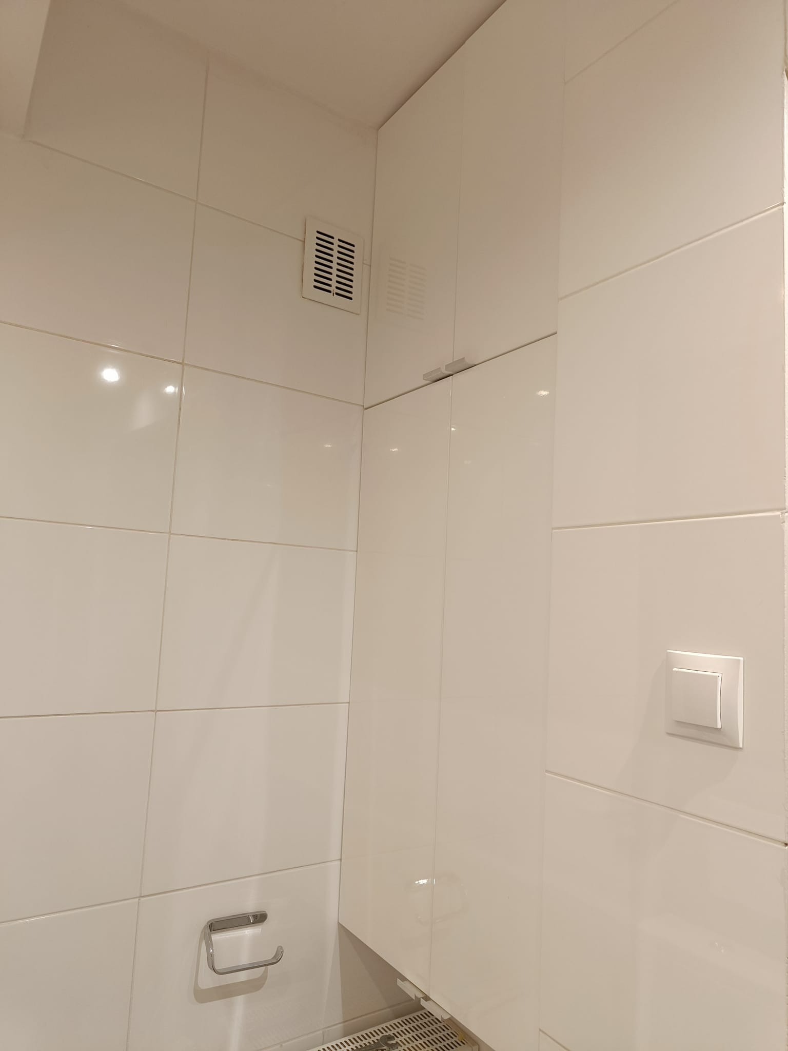 Biała szafka wisząca z połyskiem, wbudowana w ścianę z białych płytek, widoczny włącznik światła i kratka wentylacyjna