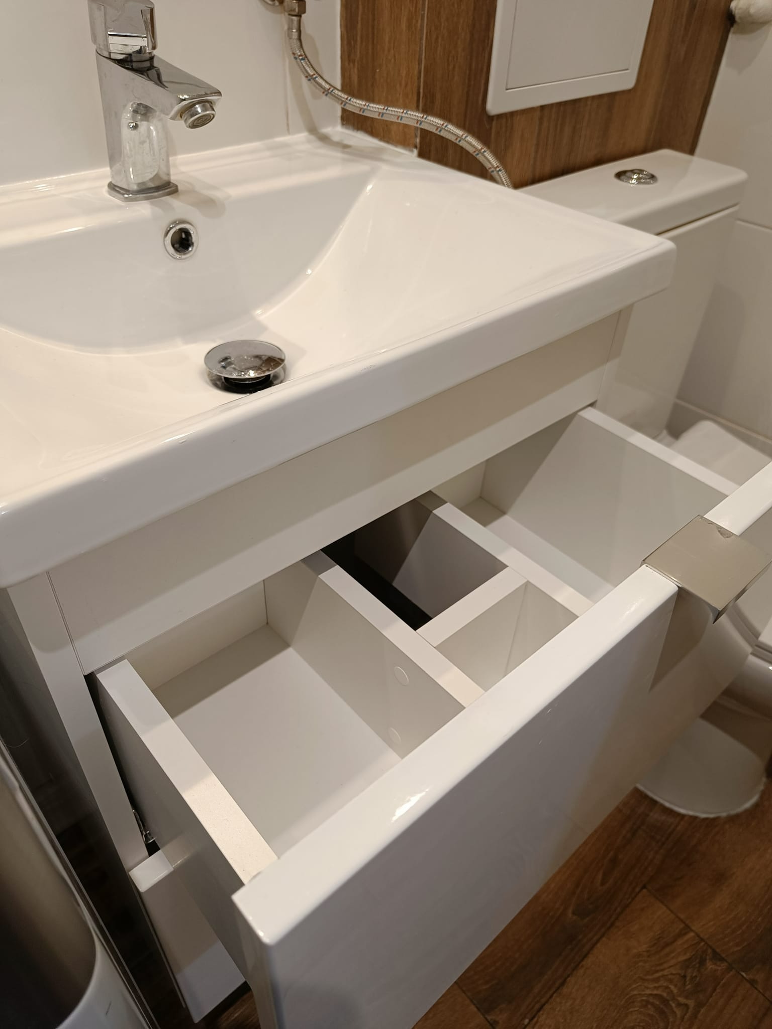 Biała umywalka z otwartą szafką pod spodem, ukazującą wewnętrzne przegródki szuflady. W tle widoczna toaleta i elementy instalacji wodnej.