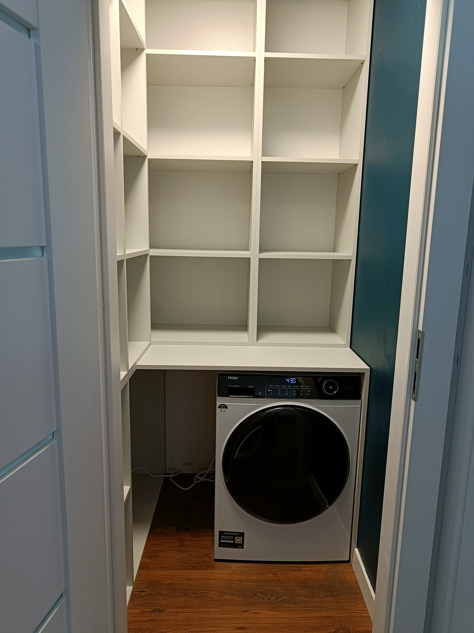 Biała szafa wnękowa z półkami, zintegrowana z pralko-suszarką Haier, widoczna we Wrocławiu. Półki puste, wnętrze oświetlone.