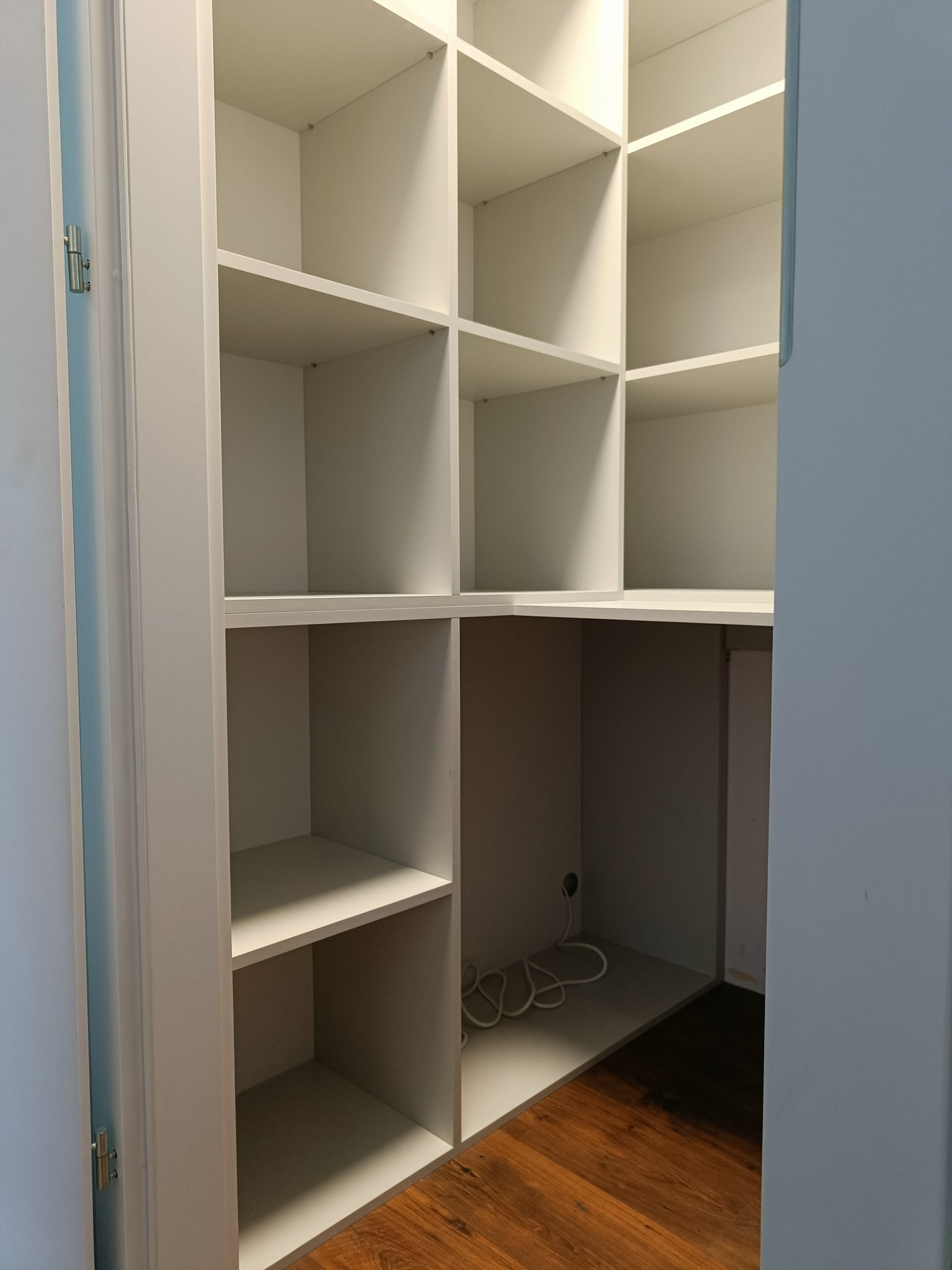 Wnętrze białej, narożnej garderoby z półkami, widoczne zawiasy drzwi i kabel zasilający w dolnej części.