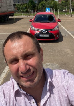 Autoportret mężczyzny na placu manewrowym z czerwonym samochodem osobowym z literą L na dachu w tle. Widoczny numer rejestracyjny pojazdu.