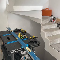 Adrian Michalski - Wnętrze w trakcie remontu: białe schody z narzędziami na blacie roboczym, poziomica i skrzynka narzędziowa w tle. Widok na surowe schody w trakcie wykończenia.