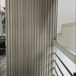 Adrian Michalski - Dekoracyjna ścianka działowa z drewnianych lameli w odcieniach szarości, oddzielająca przestrzeń, widoczna z perspektywy schodów z metalową balustradą.