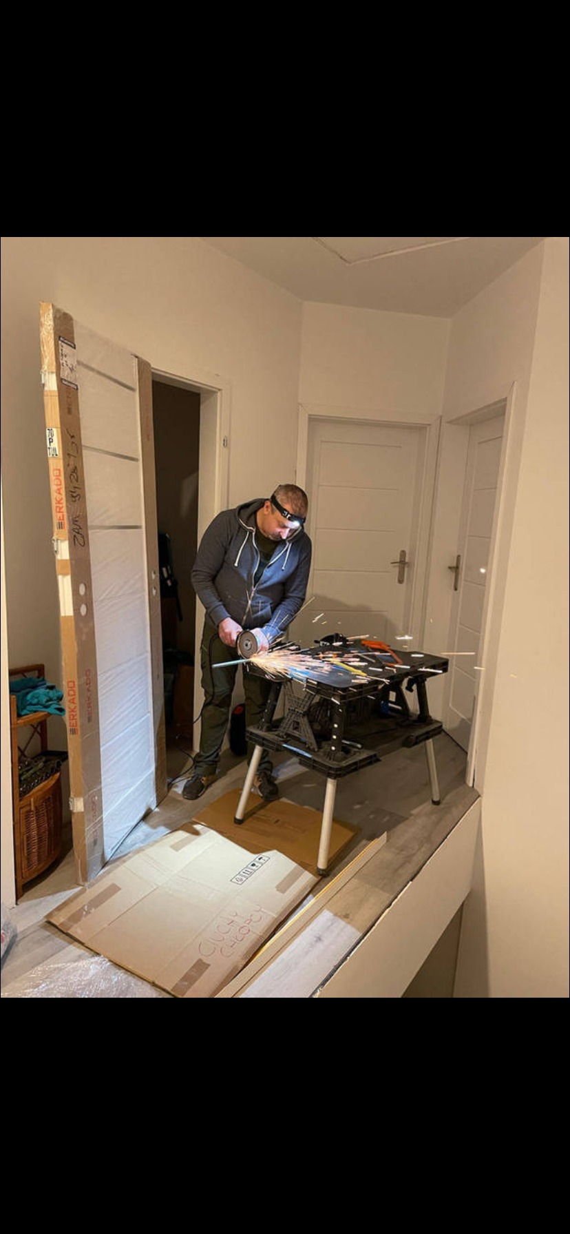 Mężczyzna w okularach ochronnych tnie metalową rurkę szlifierką kątową w korytarzu. Obok oparta o ścianę nowa, zapakowana ościeżnica.