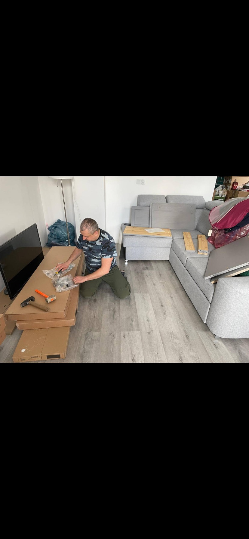 Mężczyzna w pokoju na kolanach skręca meble. Obok rozłożone elementy sofy, narzędzia i kartony. W tle telewizor i podłoga z paneli.