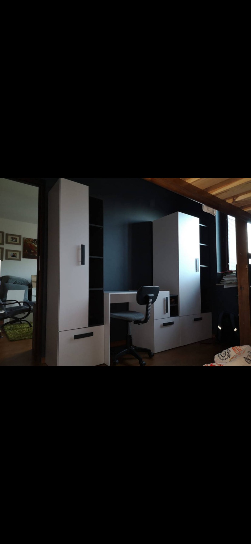 Nowoczesny pokój z białymi meblami modułowymi i biurkiem, czarne akcenty. Krzesło biurowe na kółkach. Widok na inny pokój przez otwarte drzwi.