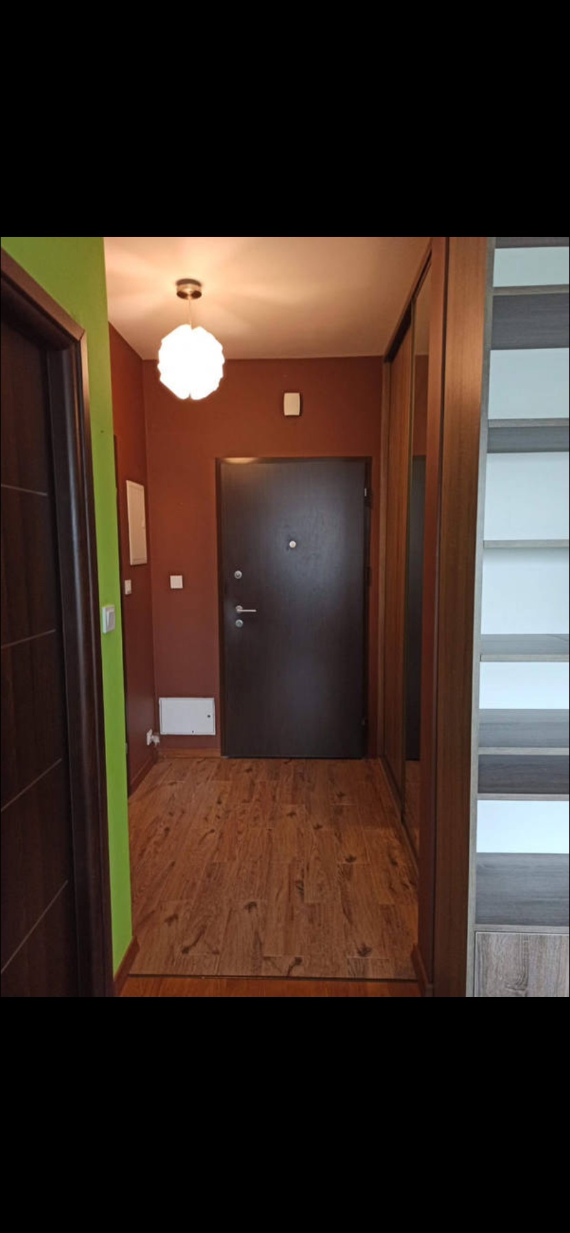 Wykończony przedpokój: brązowe ściany, drewniana podłoga, drzwi wejściowe i szafa wnękowa. Akcent kolorystyczny: zielona ściana przy wejściu do innego pomieszczenia.