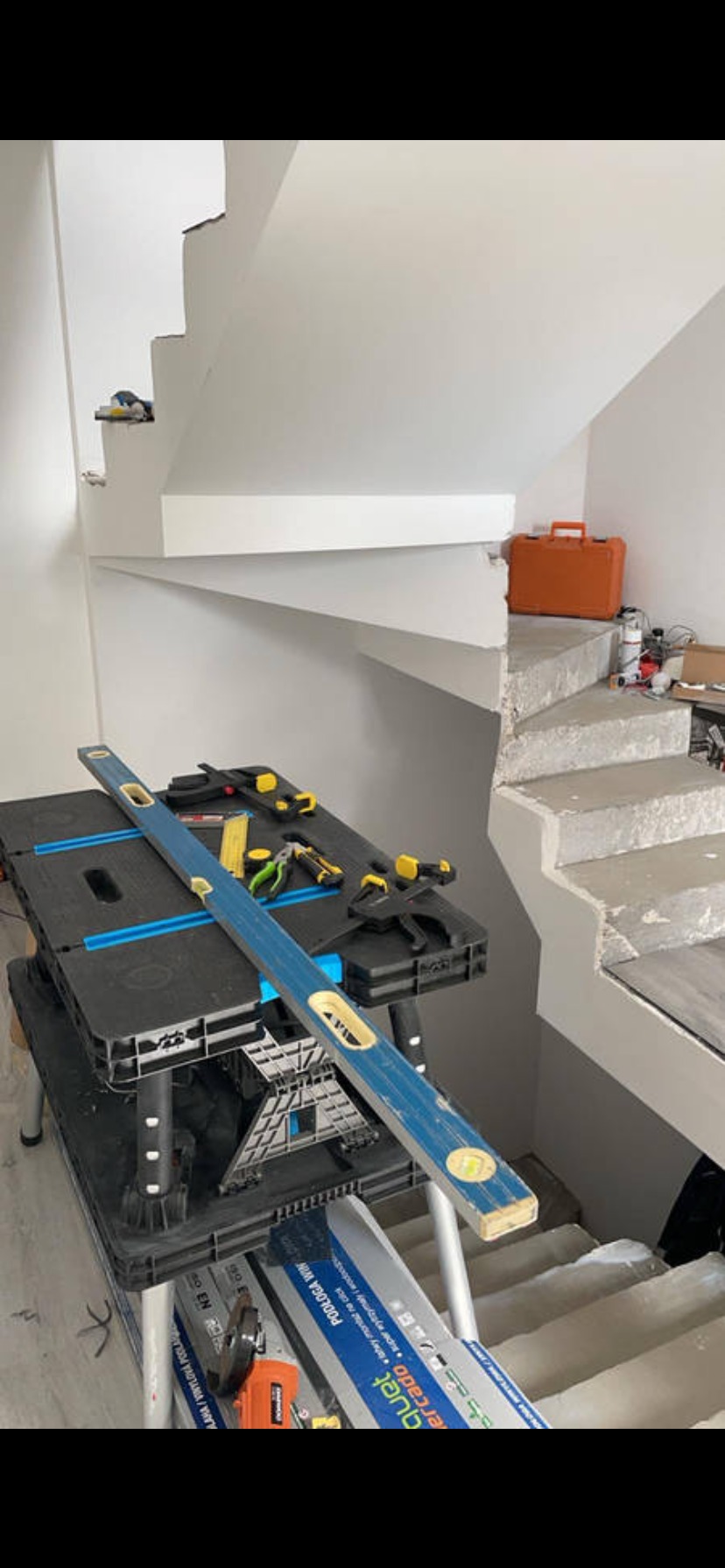 Wnętrze w trakcie remontu: białe schody z narzędziami na blacie roboczym, poziomica i skrzynka narzędziowa w tle. Widok na surowe schody w trakcie wykończenia.