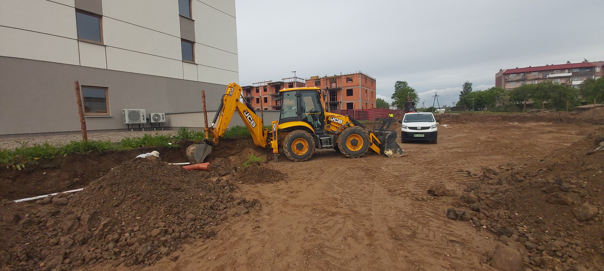 Żółta koparko-ładowarka JCB przy wykopie z rurami na placu budowy; w tle budynek w trakcie budowy i biały samochód dostawczy.