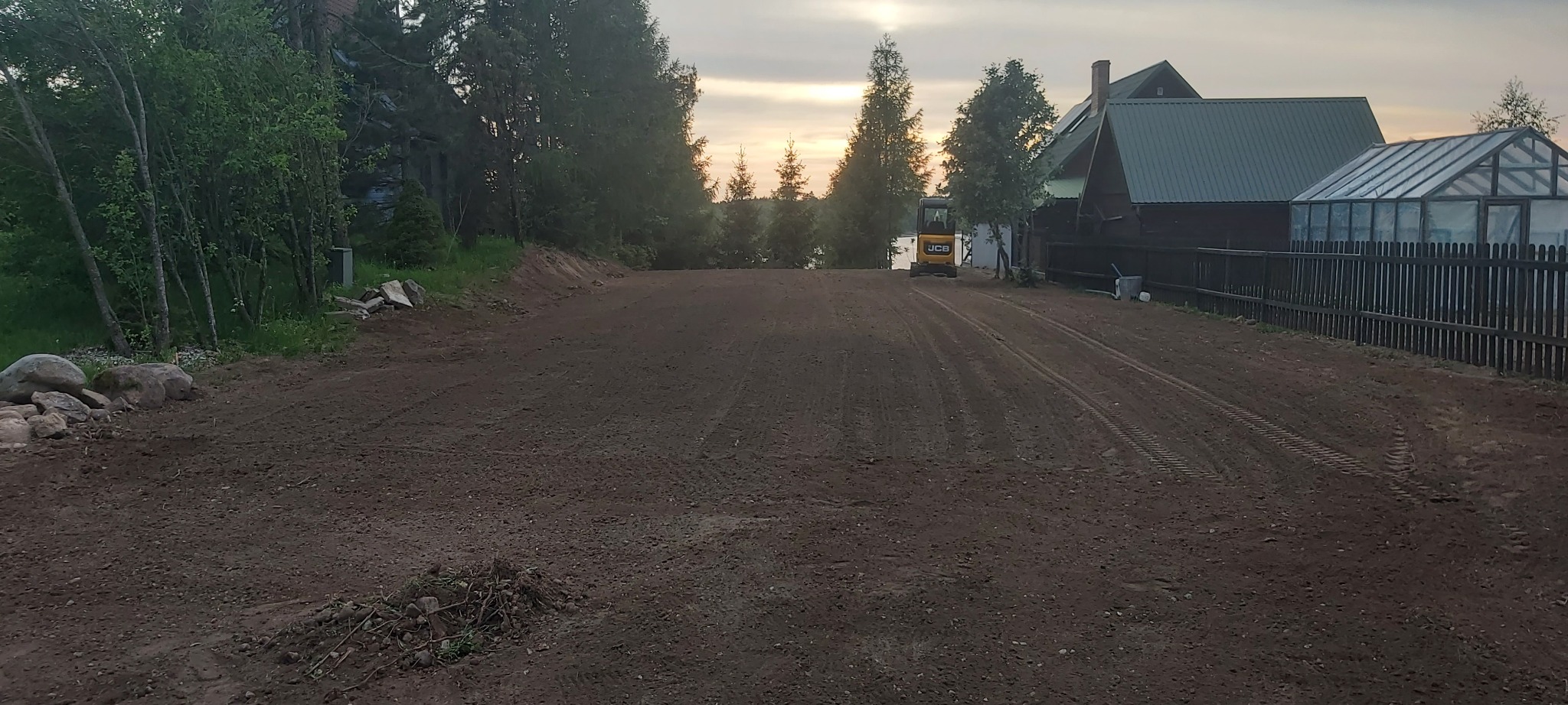 Żółta mini koparka JCB na wyrównanym, przygotowanym terenie pod budowę, widoczne ślady opon, w tle domy i drzewa, wieczorne oświetlenie.