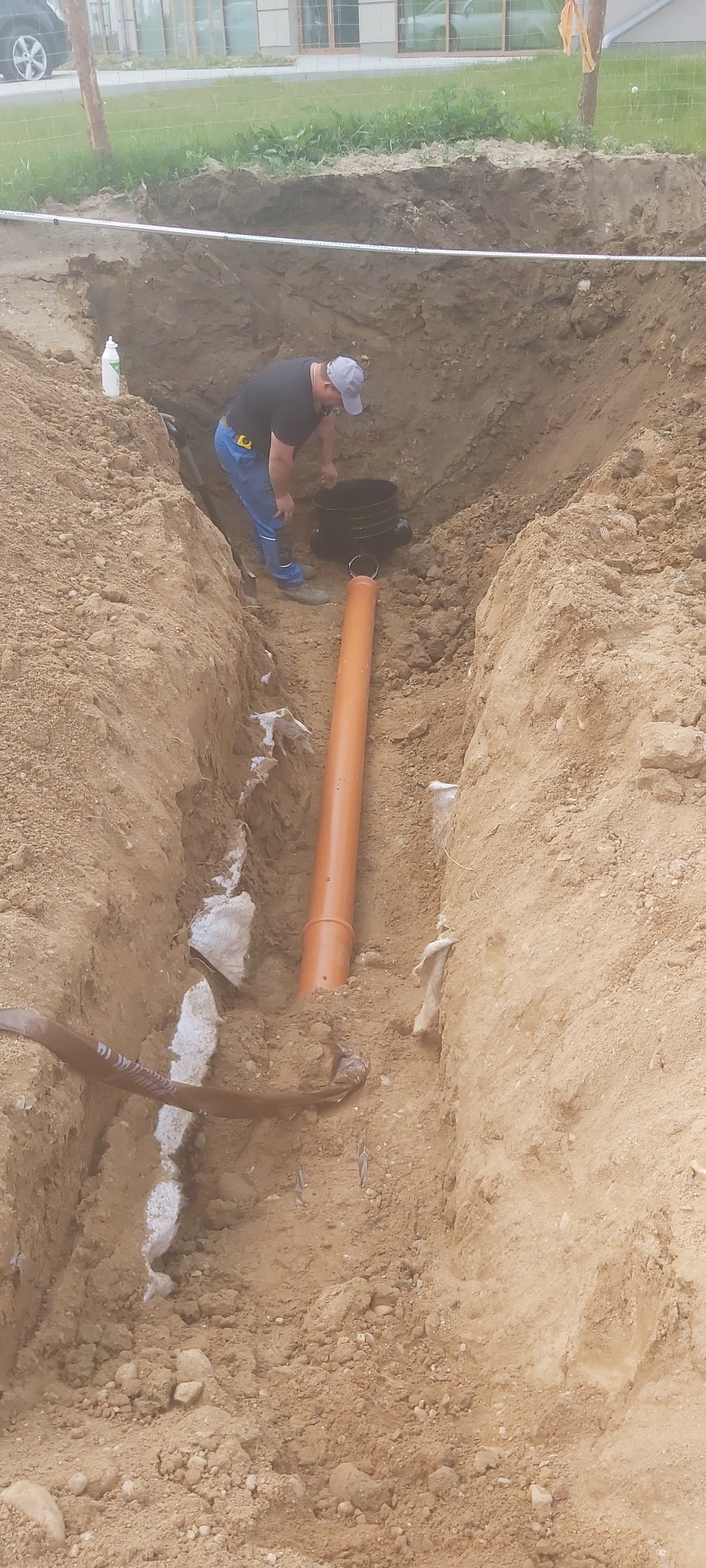 Pracownik instaluje rurę kanalizacyjną w wykopie o ziemnych ścianach, widoczne elementy instalacji i narzędzia. Prace ziemne w toku.