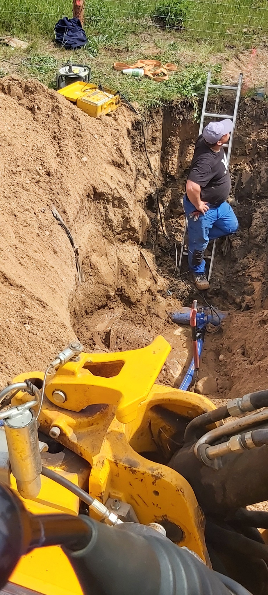 Głęboki wykop ziemny z robotnikiem na drabinie, widoczne rury instalacji i fragment maszyny budowlanej. Prace ziemne w trakcie realizacji.