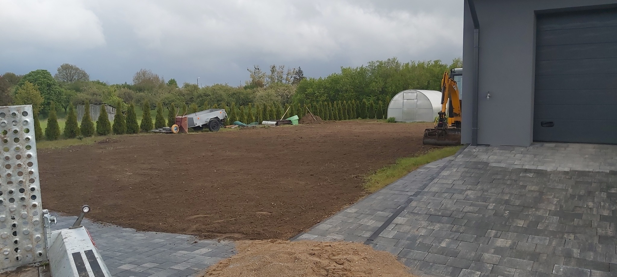 Przygotowany teren pod budowę z traktorem, przyczepką i stertą piasku. Widoczna kostka brukowa, budynek z garażem i szpaler drzew. Pochmurne niebo.