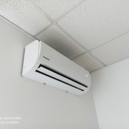 Optima Klima - Biały klimatyzator marki Kaisai zamontowany na jasnej ścianie w pomieszczeniu z kasetonowym sufitem.