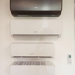 Optima Klima - Cztery klimatyzatory marki Gree zamontowane na białej ścianie, w tym jeden w kolorze grafitowym, z widocznym pilotem po prawej stronie.
