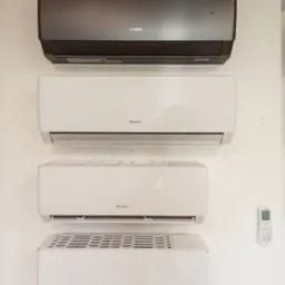 Cztery klimatyzatory marki Gree zamontowane na białej ścianie, w tym jeden w kolorze grafitowym, z widocznym pilotem po prawej stronie.