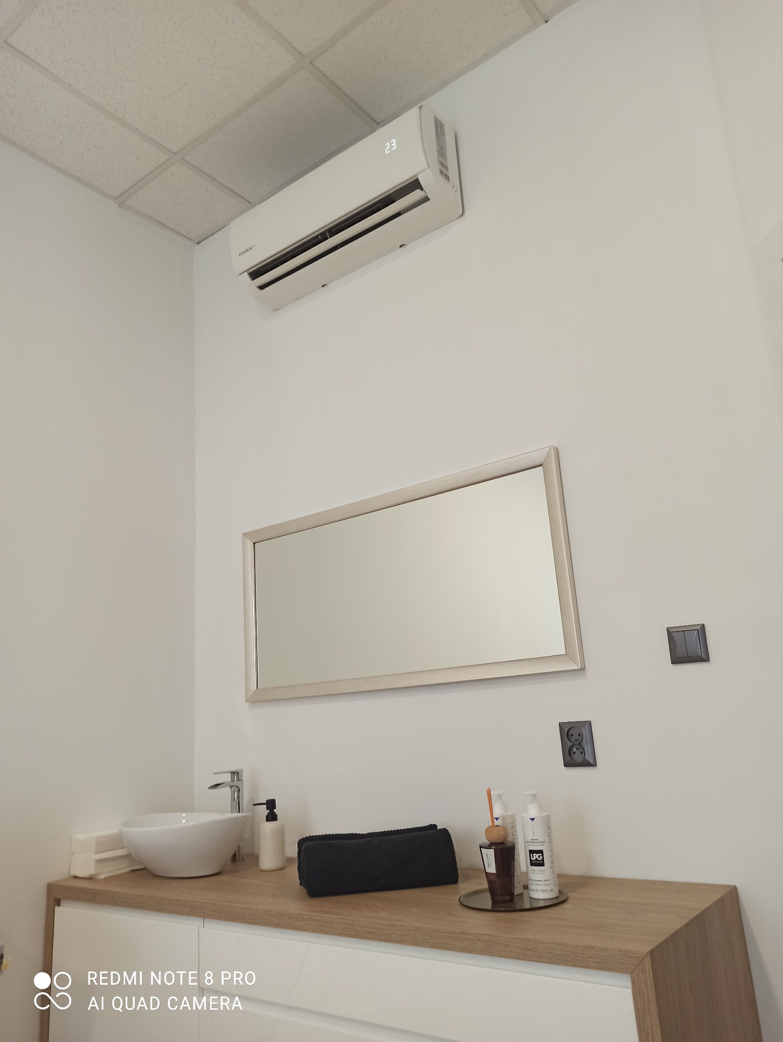 Biały klimatyzator ścienny z wyświetlaczem temperatury 23 stopnie, zainstalowany nad umywalką w łazience z lustrem i akcesoriami.