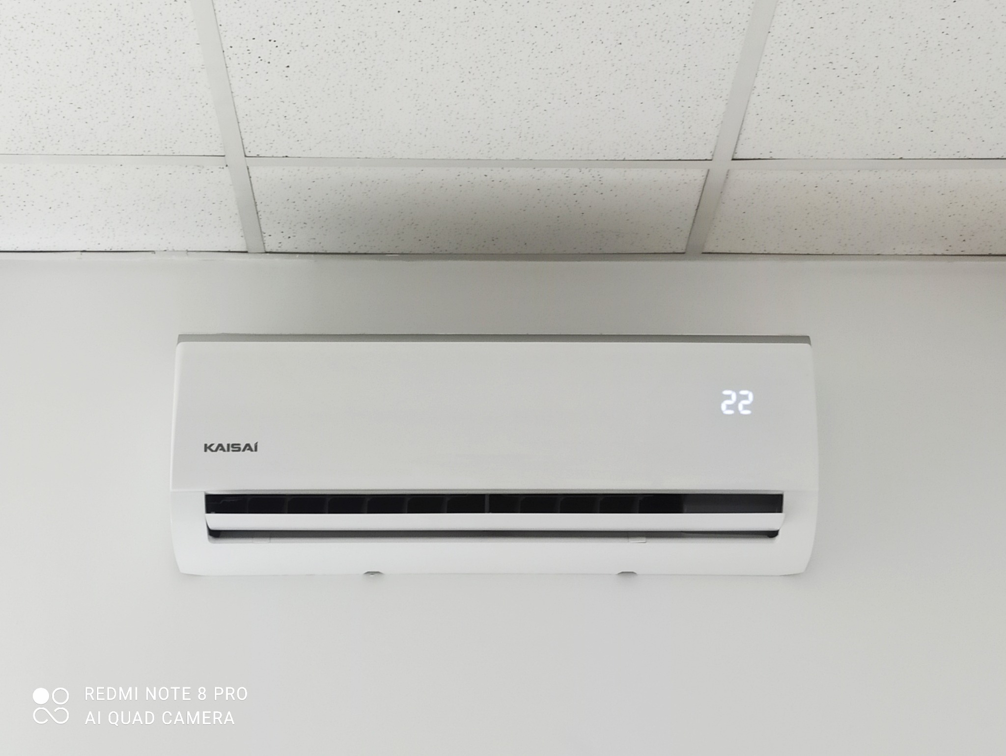 Biały klimatyzator Kaisai z wyświetlaną temperaturą 22 stopnie, zamontowany na białej ścianie pod kasetonowym sufitem.