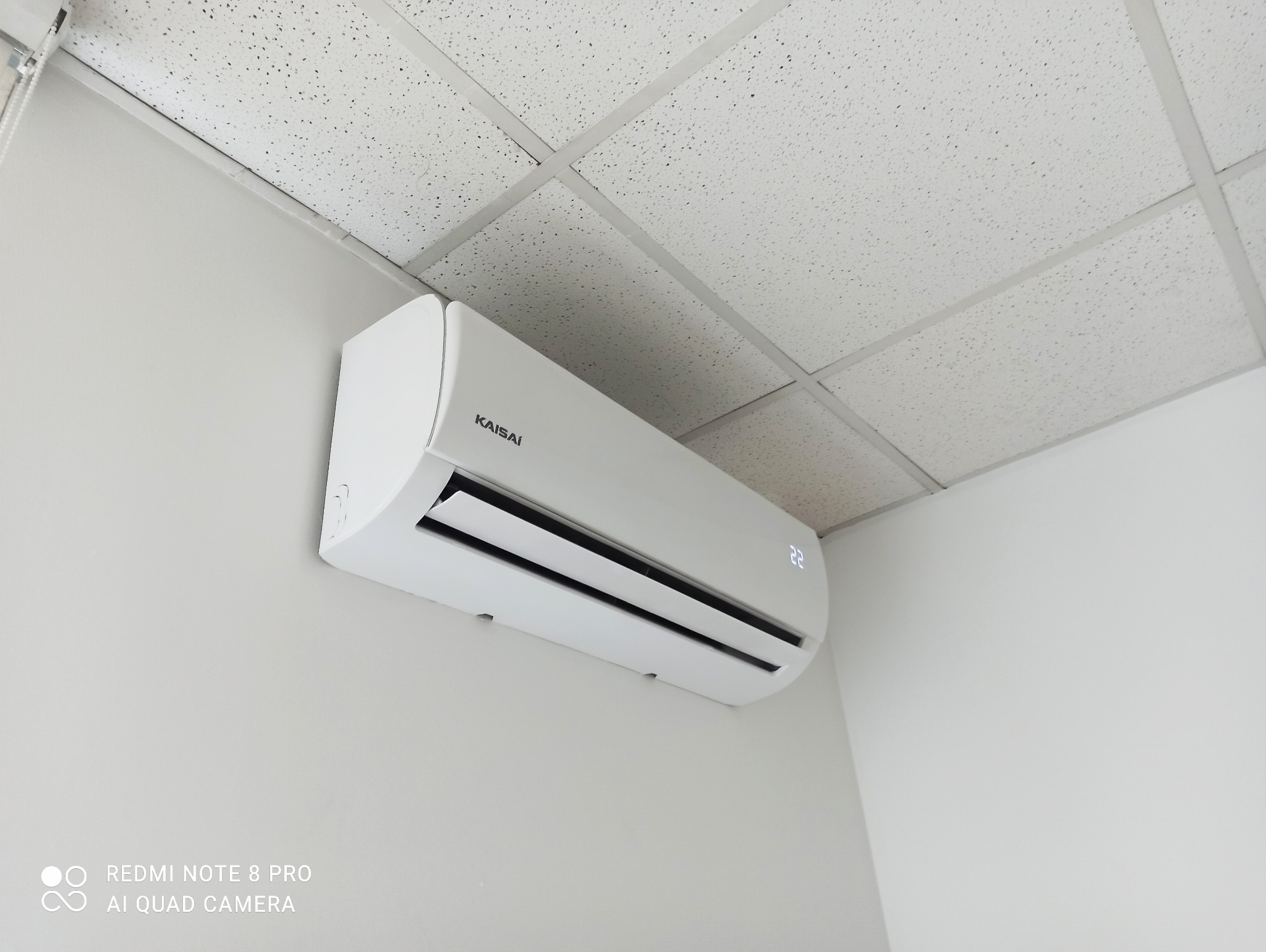 Biały klimatyzator marki Kaisai zamontowany na jasnej ścianie w pomieszczeniu z kasetonowym sufitem.