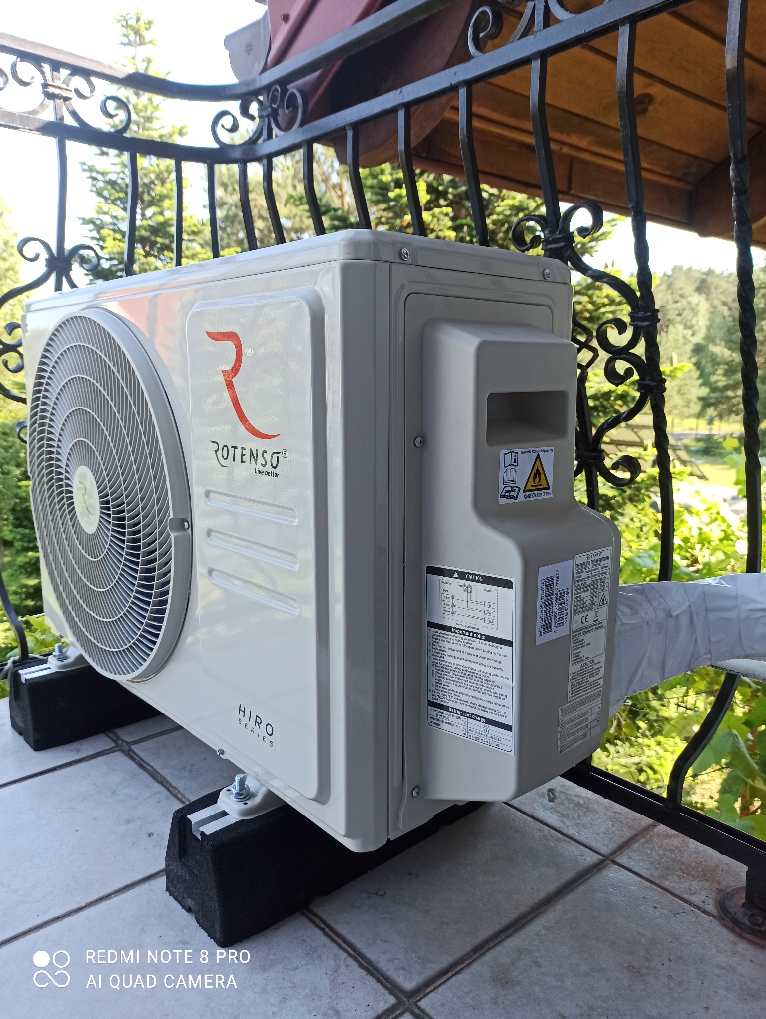 Zewnętrzna jednostka klimatyzacji Rotenso Hiro Series zamontowana na balkonie z kutą balustradą, widoczne etykiety informacyjne i logo producenta.