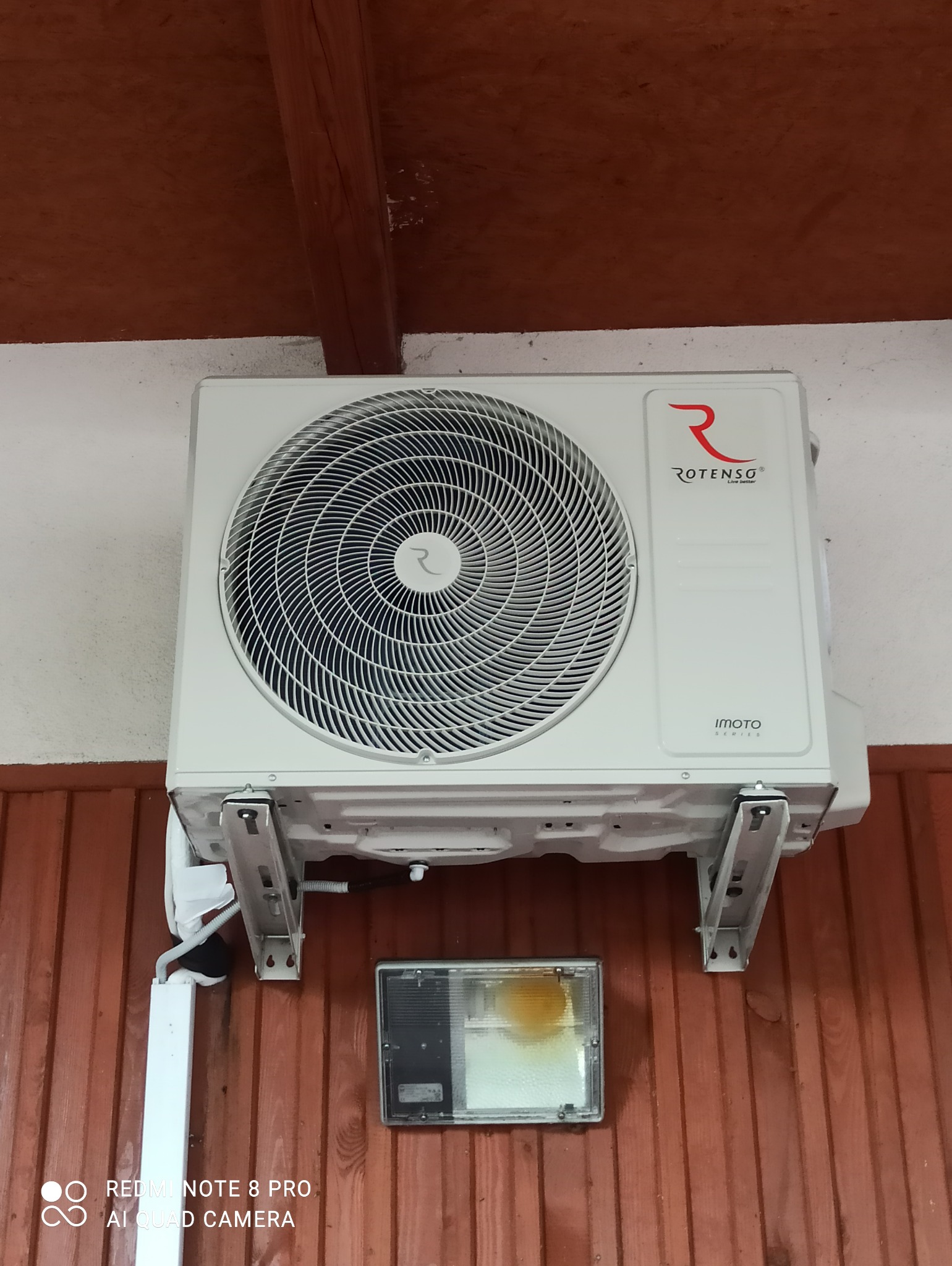 Biała jednostka zewnętrzna klimatyzatora Rotenso zamontowana na metalowych wspornikach na brązowej, drewnianej ścianie. Widoczny fragment białej rury i lampa halogenowa.