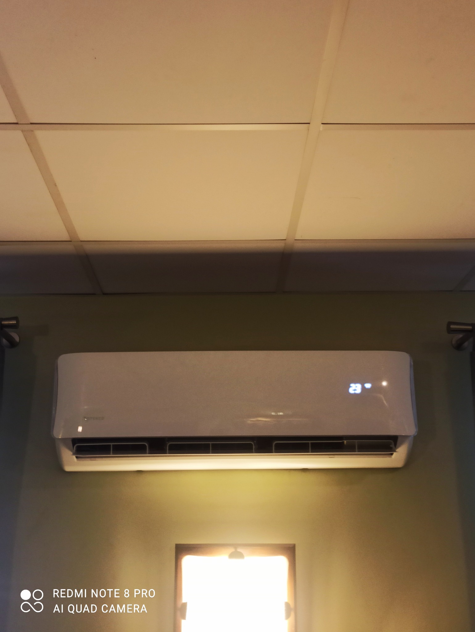 Biała jednostka klimatyzacji ściennej z wyświetlaczem temperatury 23 stopnie Celsjusza, zamontowana na zielonej ścianie pod sufitem kasetonowym.