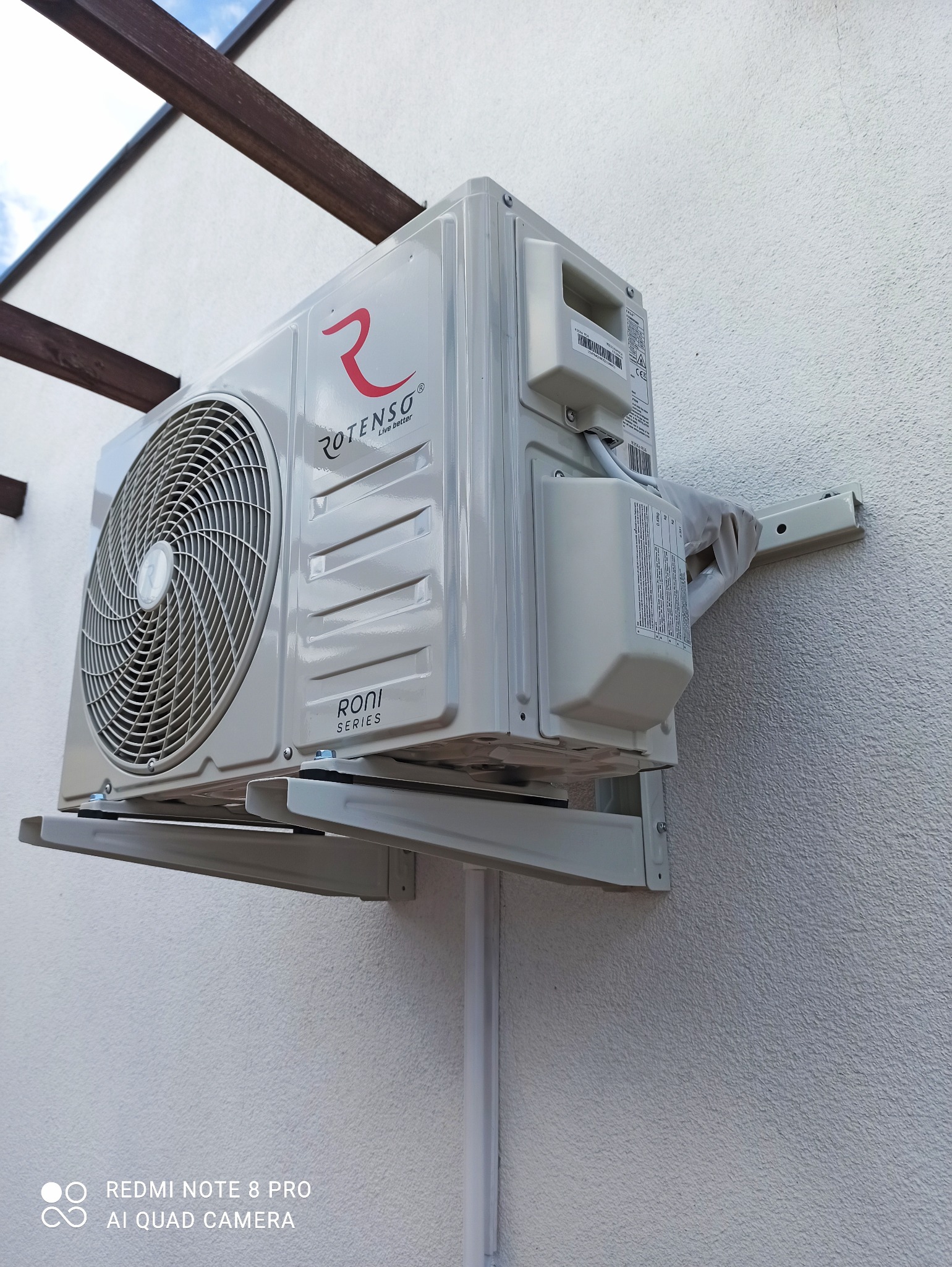 Zewnętrzna jednostka klimatyzacji Rotenso zamontowana na białej elewacji budynku z widocznymi elementami montażowymi i rurami odprowadzającymi.