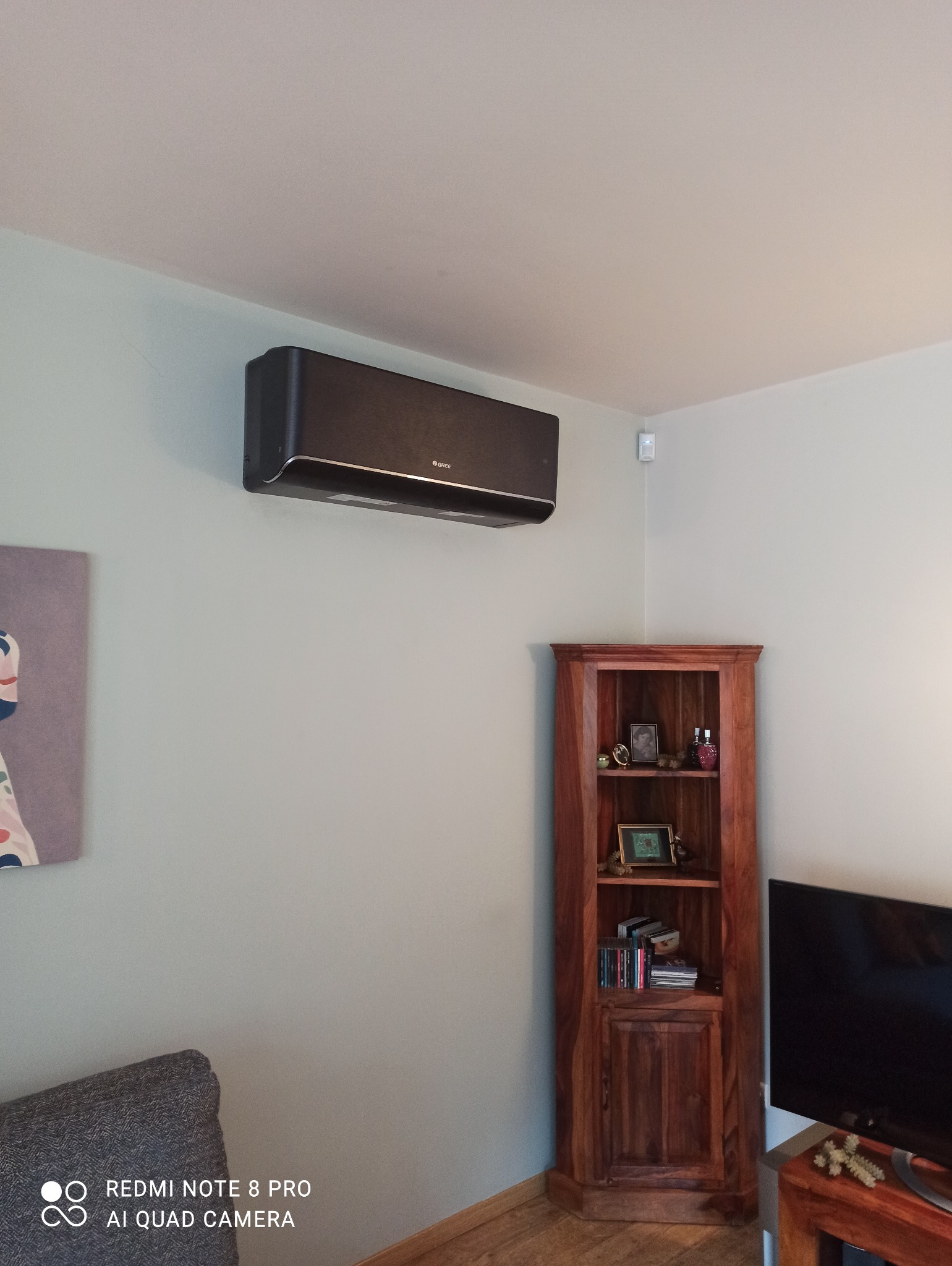 Czarny klimatyzator marki Gree zamontowany na ścianie w pokoju, obok drewnianego narożnego regału i obrazu.