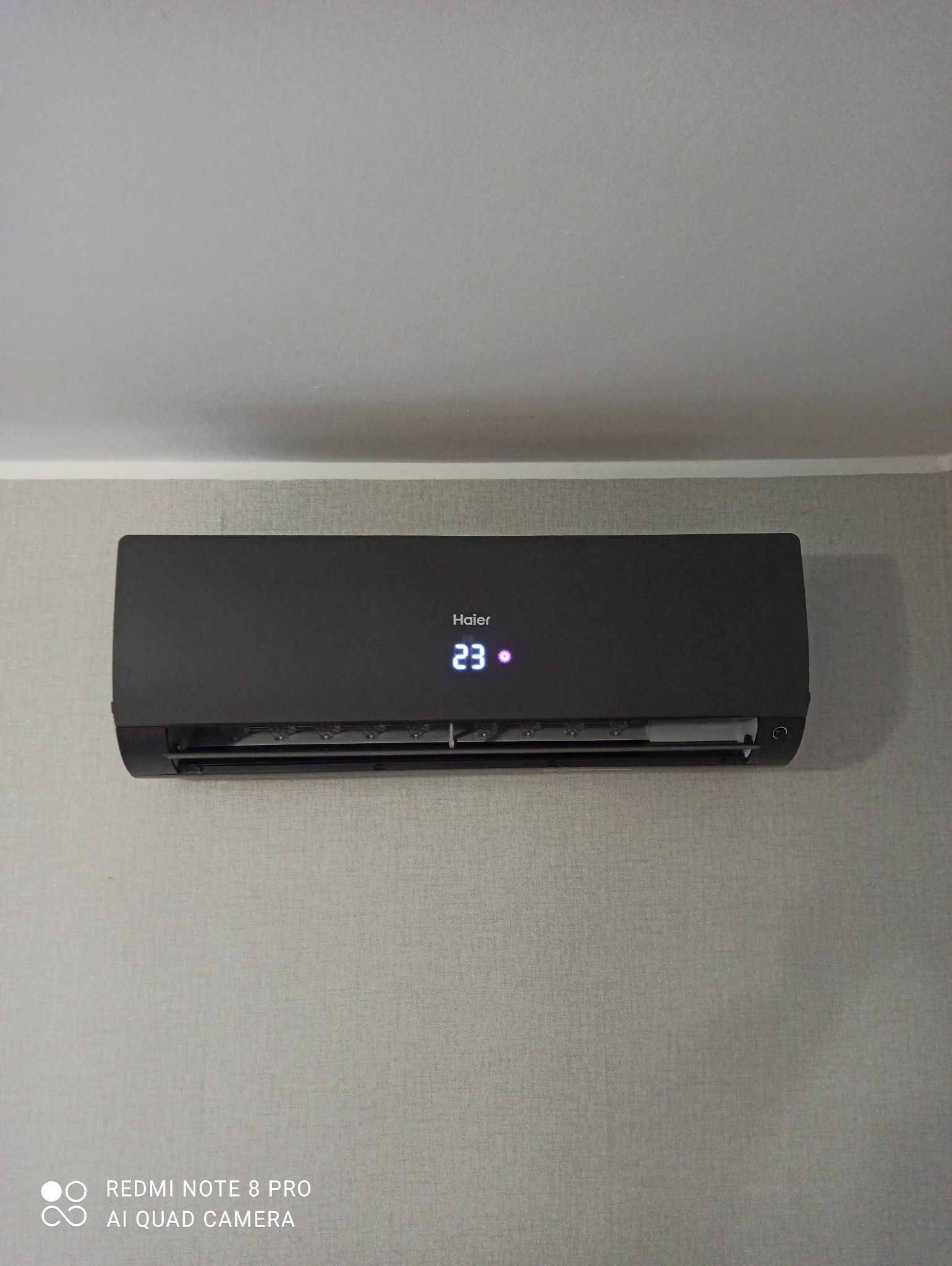 Czarny klimatyzator Haier zamontowany na szarej ścianie z wyświetlaczem wskazującym temperaturę 23 stopnie.