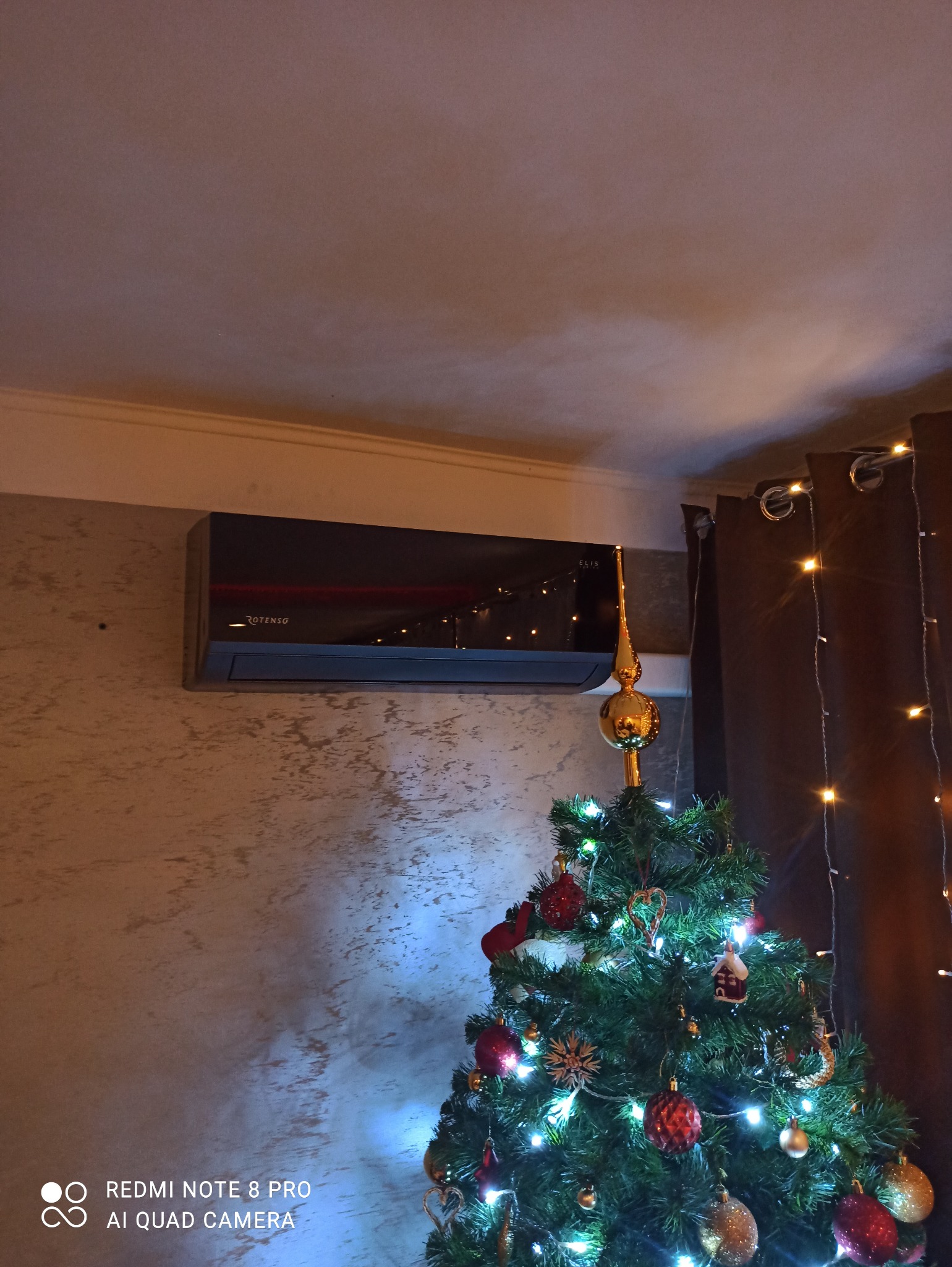 Czarny klimatyzator Rotenso Elis zamontowany na ścianie z dekoracyjną fakturą, obok choinki z lampkami i ozdobami świątecznymi.