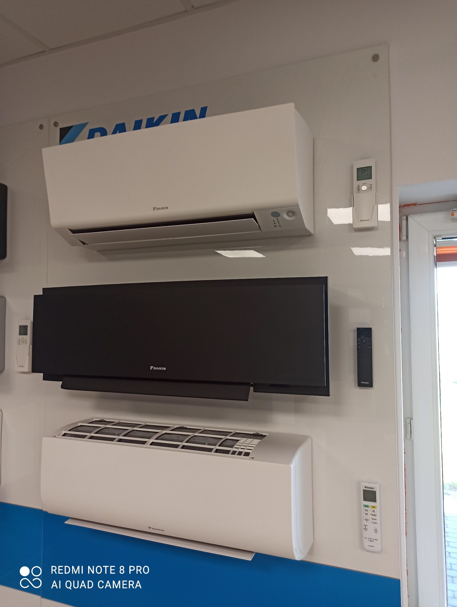 Ekspozycja trzech klimatyzatorów Daikin w kolorach białym i czarnym, wraz z dedykowanymi pilotami, prezentowanych na białym panelu z niebieskim akcentem.