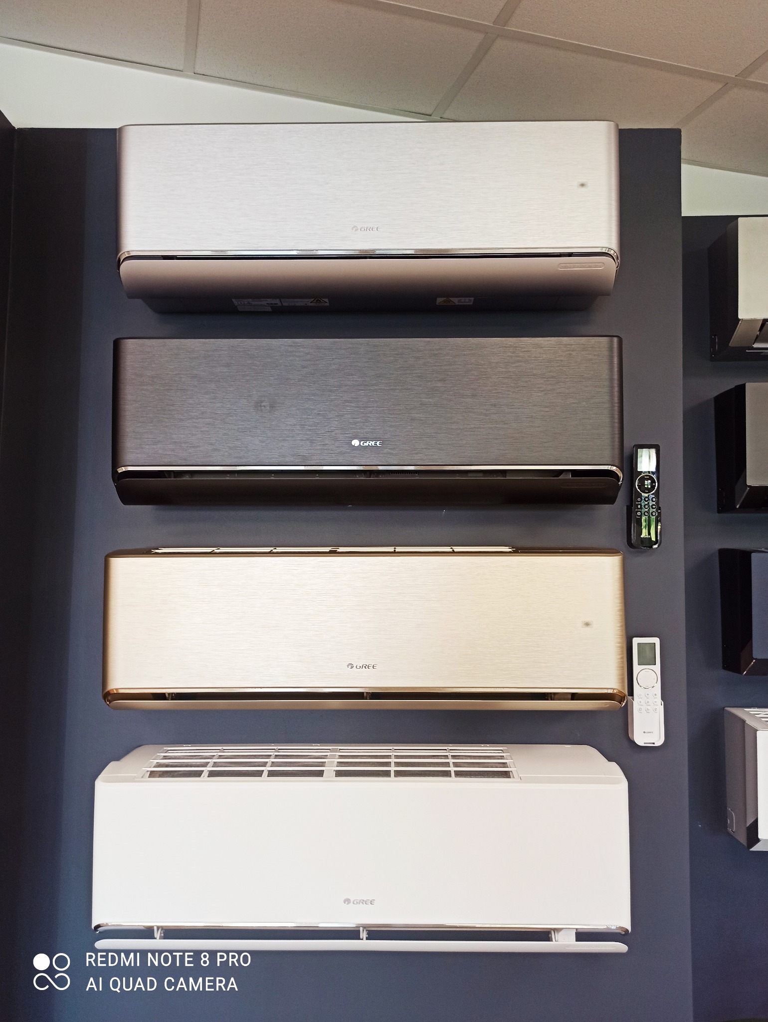 Ekspozycja czterech klimatyzatorów marki Gree w różnych kolorach (biały, złoty, szary, srebrny) zamontowanych na ciemnej ścianie, z widocznymi pilotami sterowania.