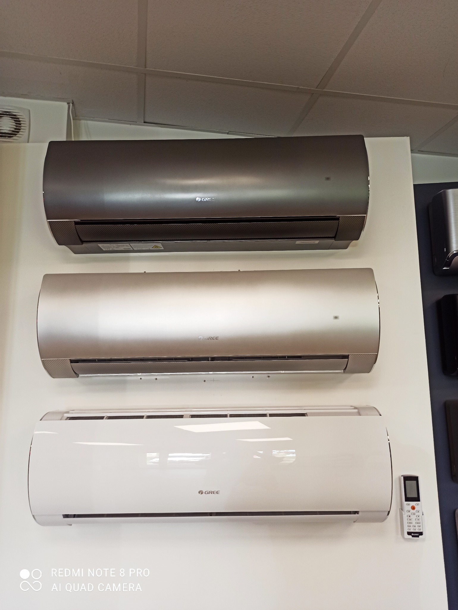 Prezentacja trzech klimatyzatorów marki Gree w różnych kolorach: grafitowym, srebrnym i białym, zamontowanych na ścianie, z pilotem do sterowania klimatyzacją na dole po prawej stronie.