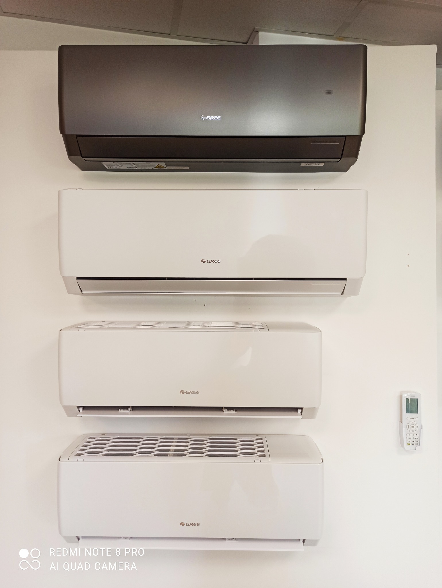 Cztery klimatyzatory marki Gree zamontowane na białej ścianie, w tym jeden w kolorze grafitowym, z widocznym pilotem po prawej stronie.