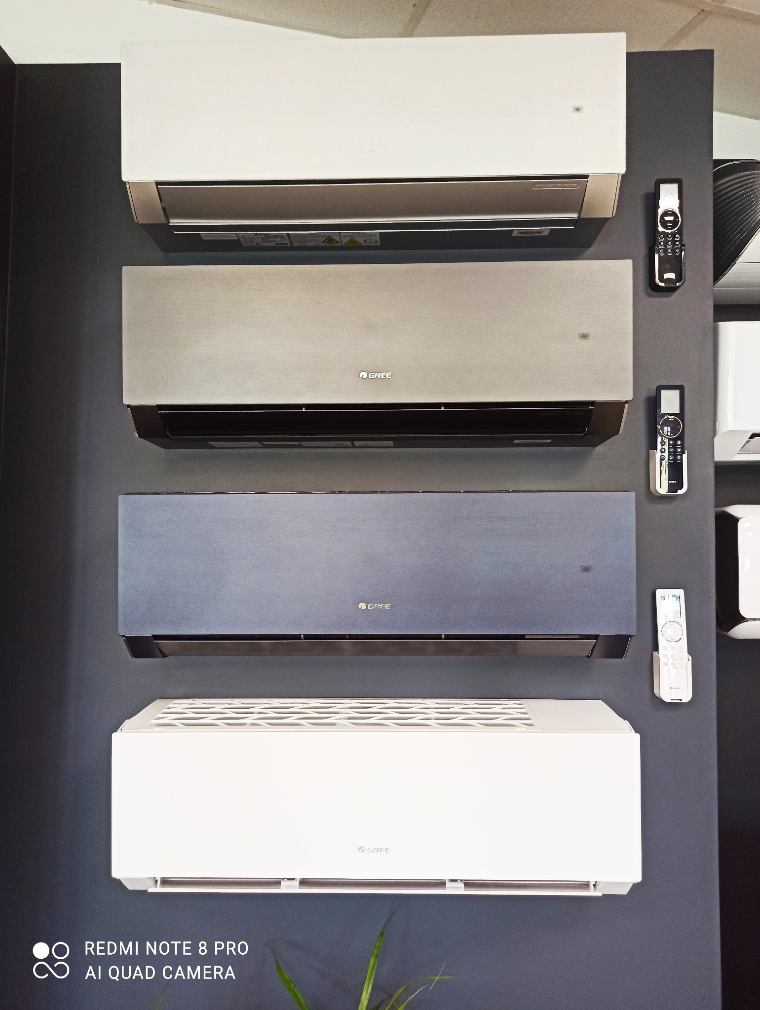 Prezentacja czterech klimatyzatorów marki Gree w różnych kolorach (biały, srebrny, szary, niebieski) zamontowanych na ciemnej ścianie, obok każdego klimatyzatora znajduje się pilot.