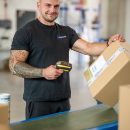 Tutenhoman Monika Homan - Pracownik magazynu skanuje kod kreskowy na kartonie przed umieszczeniem go na taśmie transportowej, widoczny tatuaż na ramieniu.