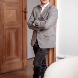 Tutenhoman Monika Homan - Uśmiechnięty mężczyzna w tweedowej marynarce, czarnych jeansach i okularach pozuje na tle drewnianych drzwi i białego fotela.