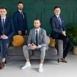 Grupa czterech mężczyzn w garniturach pozuje na szarym welurowym siedzisku przed zieloną ścianą z dekoracyjnymi panelami.