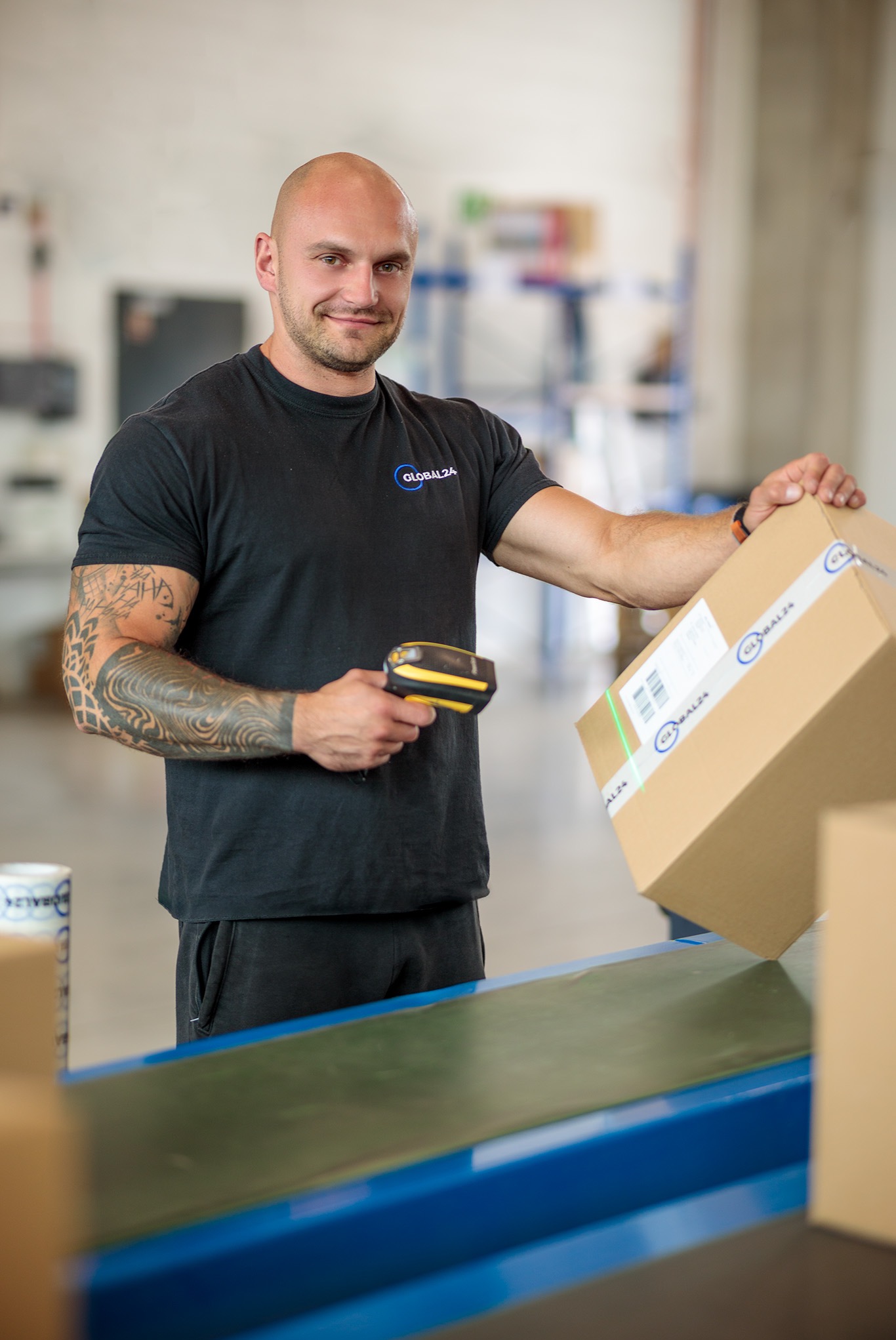 Pracownik magazynu skanuje kod kreskowy na kartonie przed umieszczeniem go na taśmie transportowej, widoczny tatuaż na ramieniu.