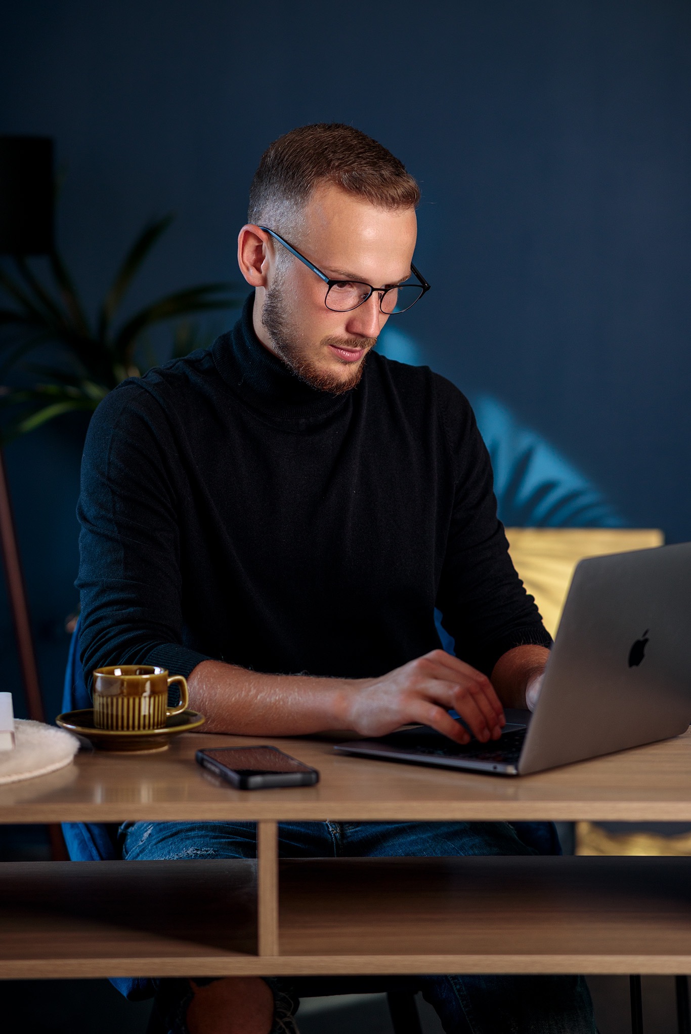 Mężczyzna w okularach i czarnym golfie pracuje na laptopie przy biurku z kawą i telefonem, w tle ciemna ściana i roślina.