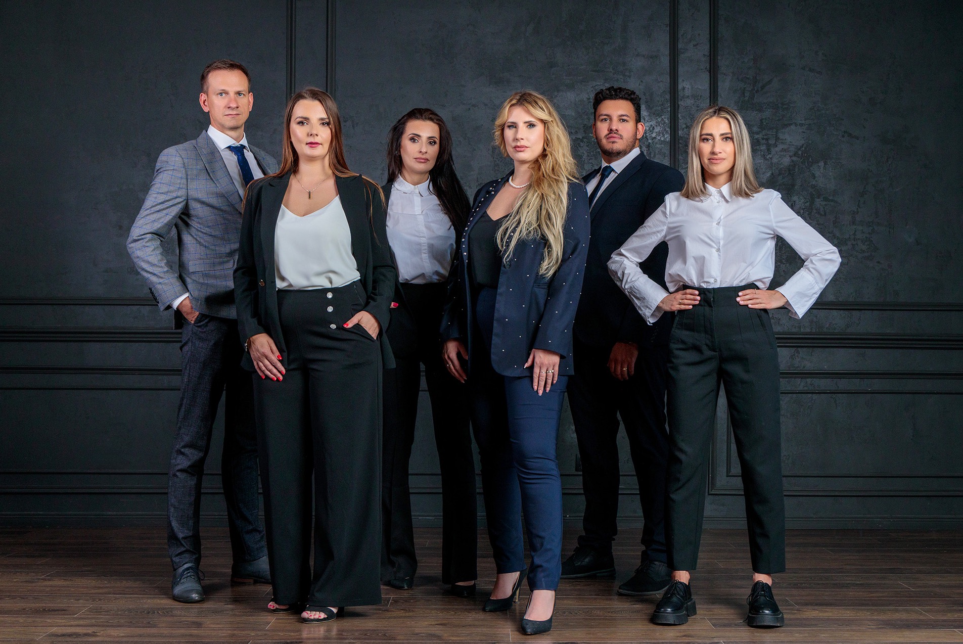 Profesjonalny portret grupowy sześciu osób w formalnych strojach na tle ciemnej ściany z panelami i drewnianej podłogi.