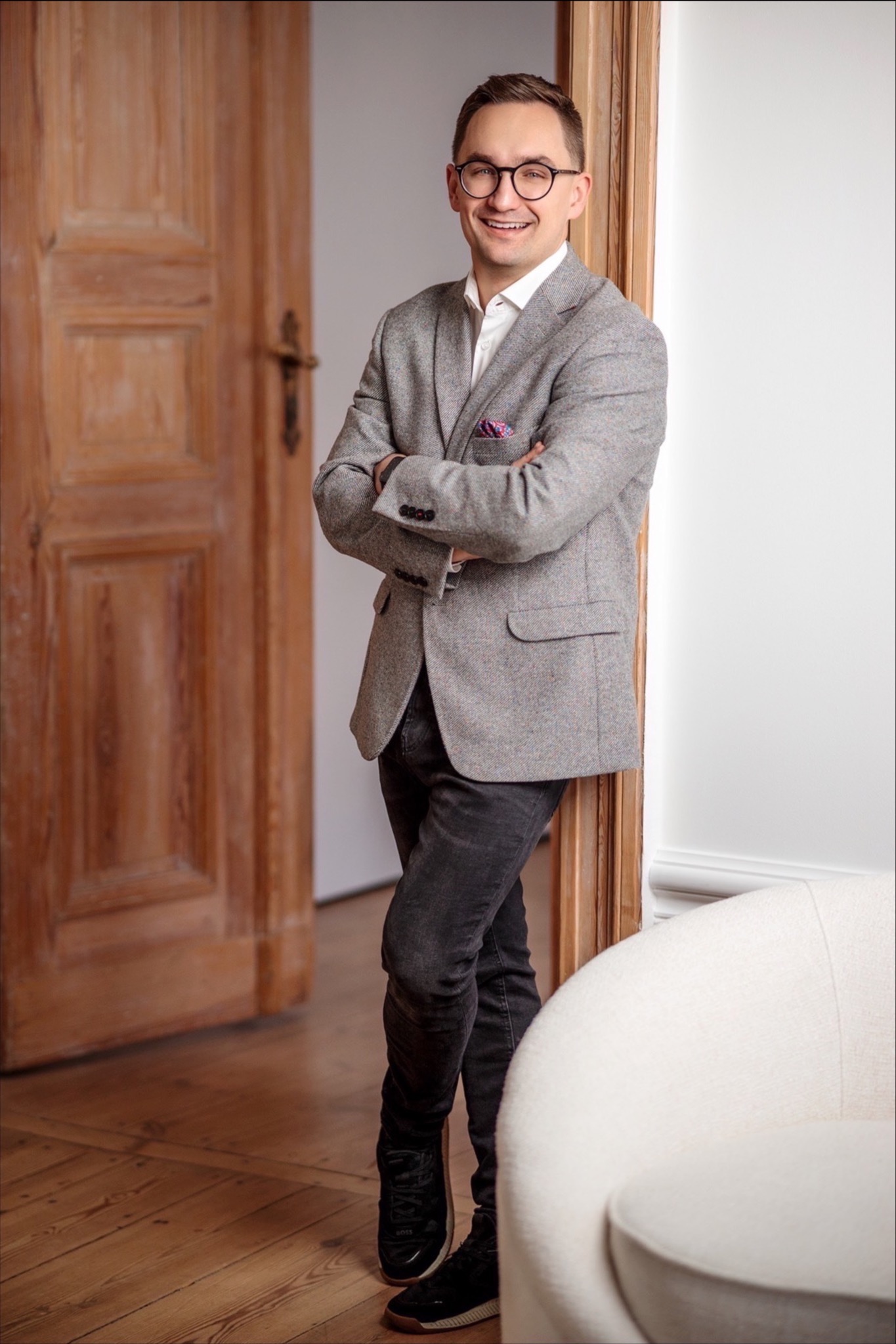 Uśmiechnięty mężczyzna w tweedowej marynarce, czarnych jeansach i okularach pozuje na tle drewnianych drzwi i białego fotela.