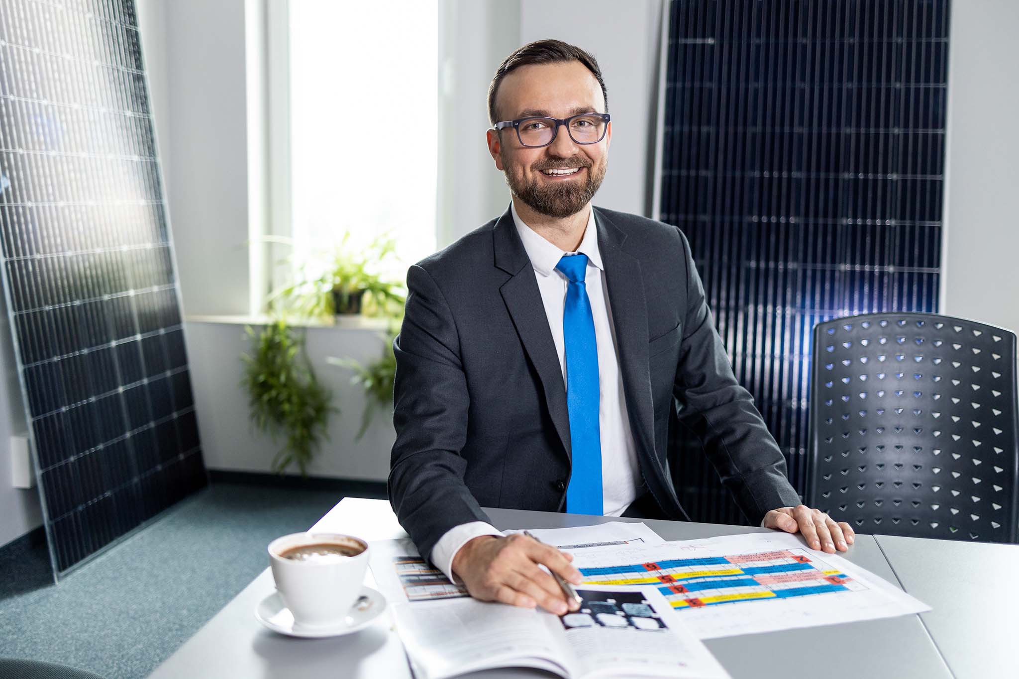 Uśmiechnięty mężczyzna w garniturze i okularach siedzi przy biurku z planami instalacji paneli słonecznych, obok filiżanka kawy, w tle panele fotowoltaiczne.