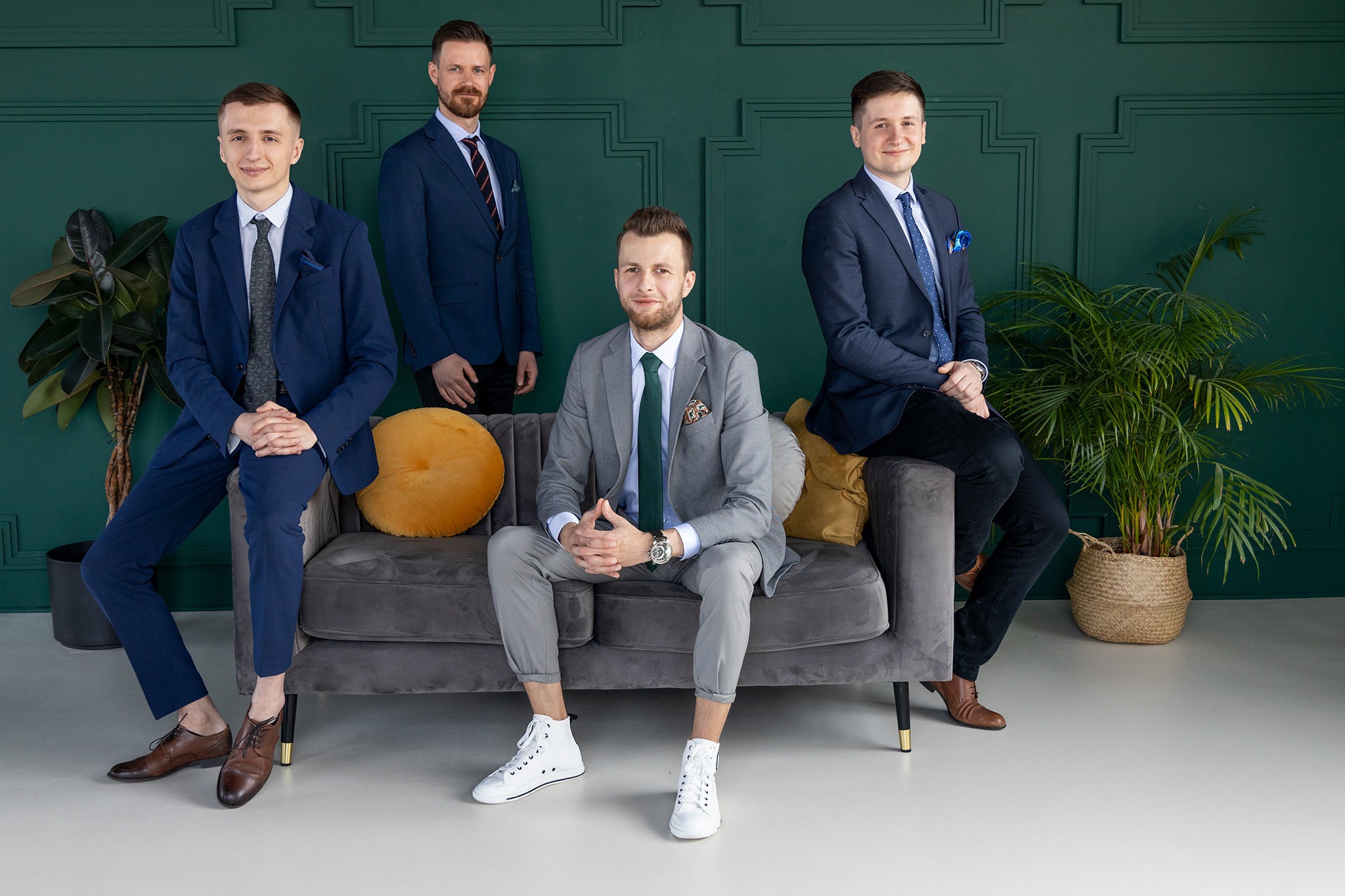 Grupa czterech mężczyzn w garniturach pozuje na szarym welurowym siedzisku przed zieloną ścianą z dekoracyjnymi panelami.