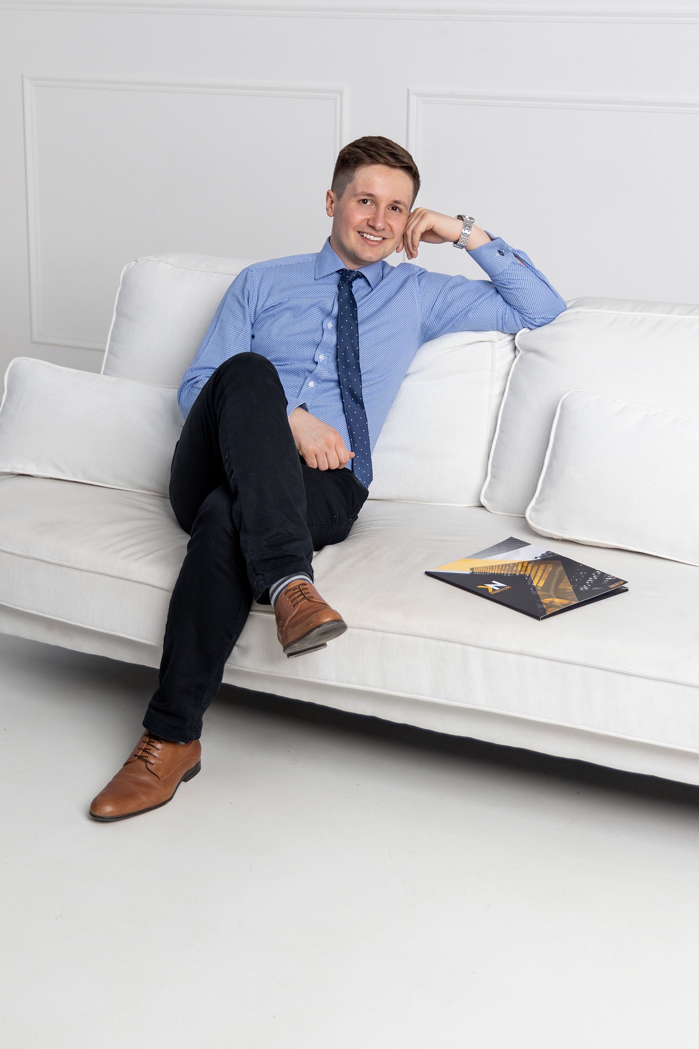 Uśmiechnięty mężczyzna w błękitnej koszuli i krawacie siedzi na białej sofie, obok leży czarna teczka z motywem architektonicznym.