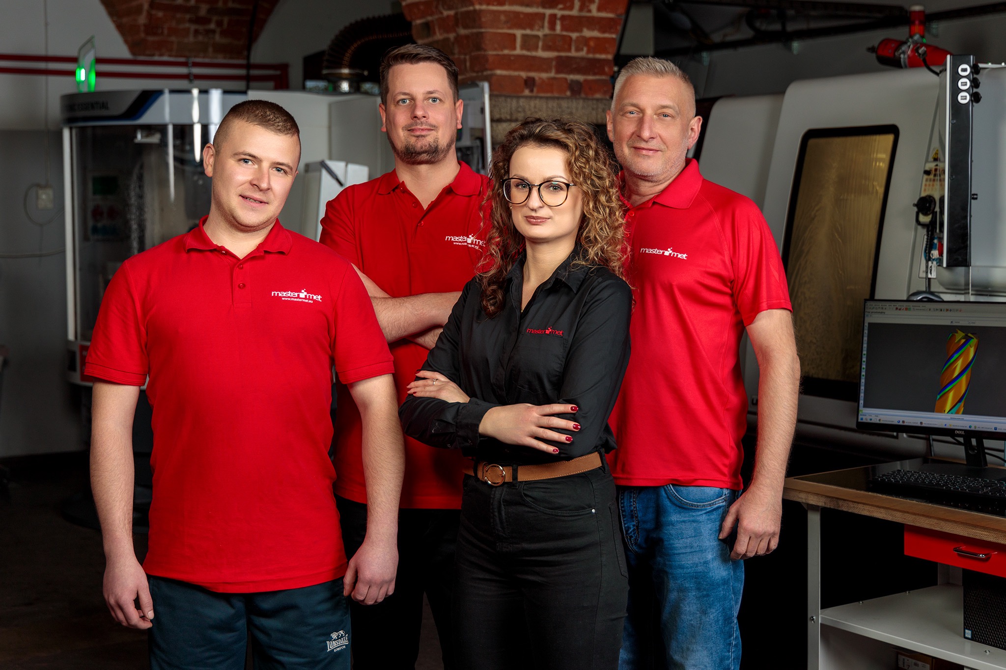 Zespół pracowników firmy Mastermet w czerwonych i czarnych koszulkach polo z logo, stojący na tle nowoczesnych maszyn CNC i ceglanej ściany.